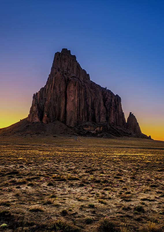 A tall rock feature rises out of the flat desert against blue sky and a sun glow on the horizon.