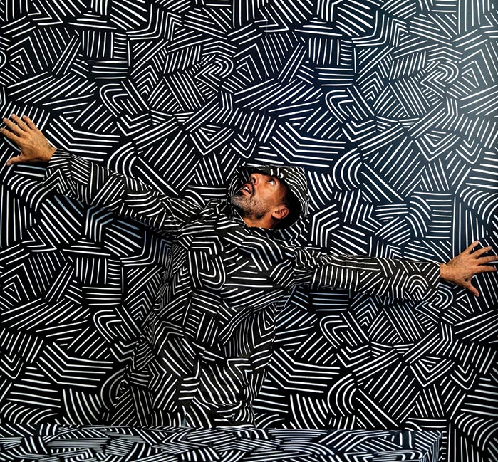 A man against a wall, where he blends in with camouflage.